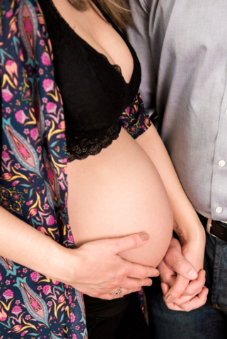 Expectant Pregnant mom wearing a black bra and floral robe holds hands with her husband as she caresses her stomach