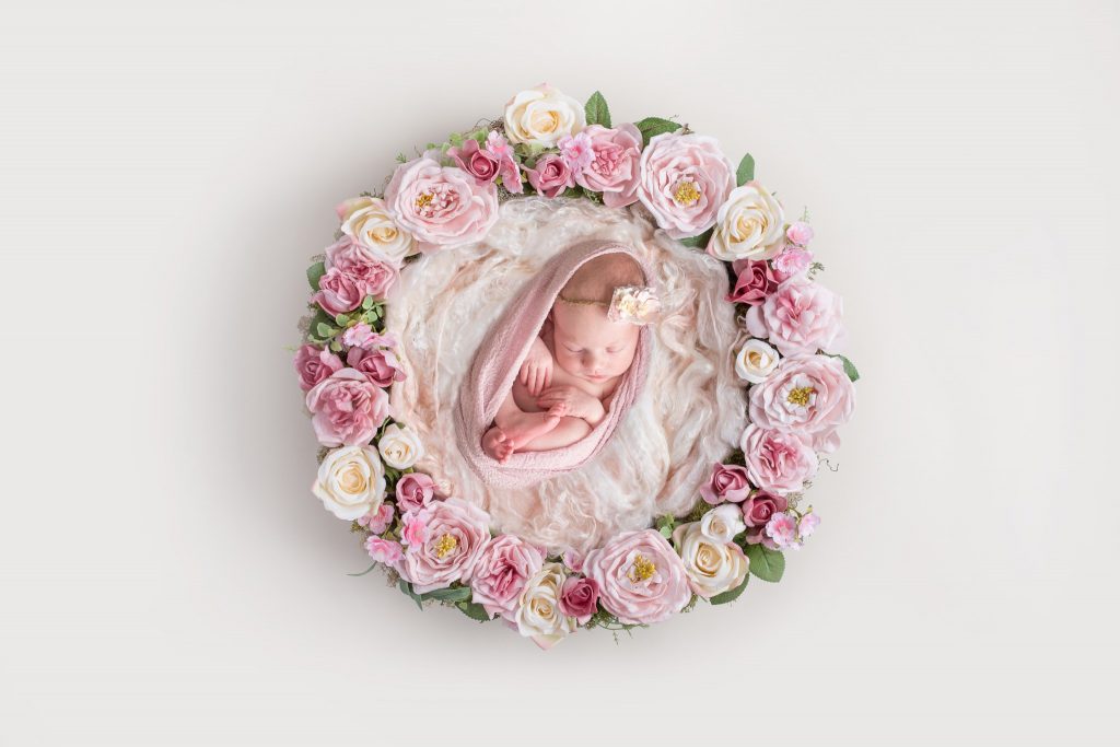 A newborn baby girl lays upon layers of pink fabric while laid in a basket adorned with florals/flowers during her newborn portrait photography session in Wexford PA near Pittsburgh