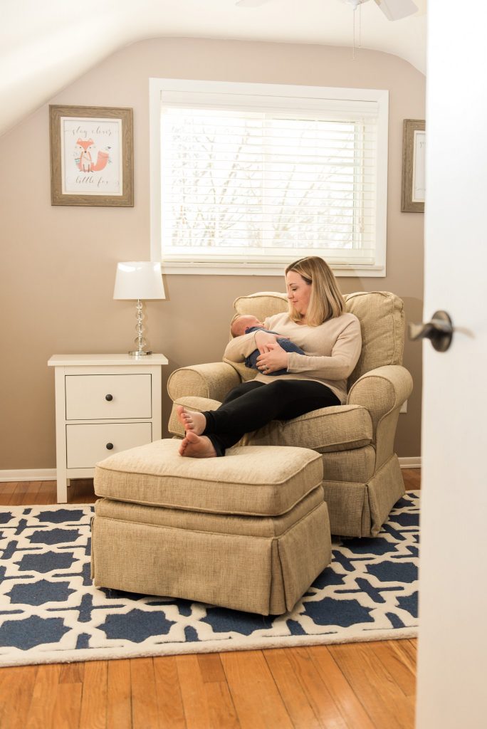 A baby boy is held in a blue swaddle while mom is seated in a neutral tan, white, and blue nursery in their Pittsburgh home.