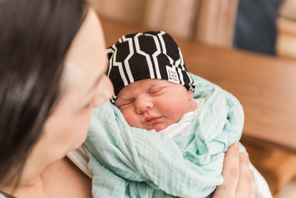 A baby boy is swaddled in a mint blanket and black beanie hat while being held by his mother for their photography session in a Pittsburgh hospital.