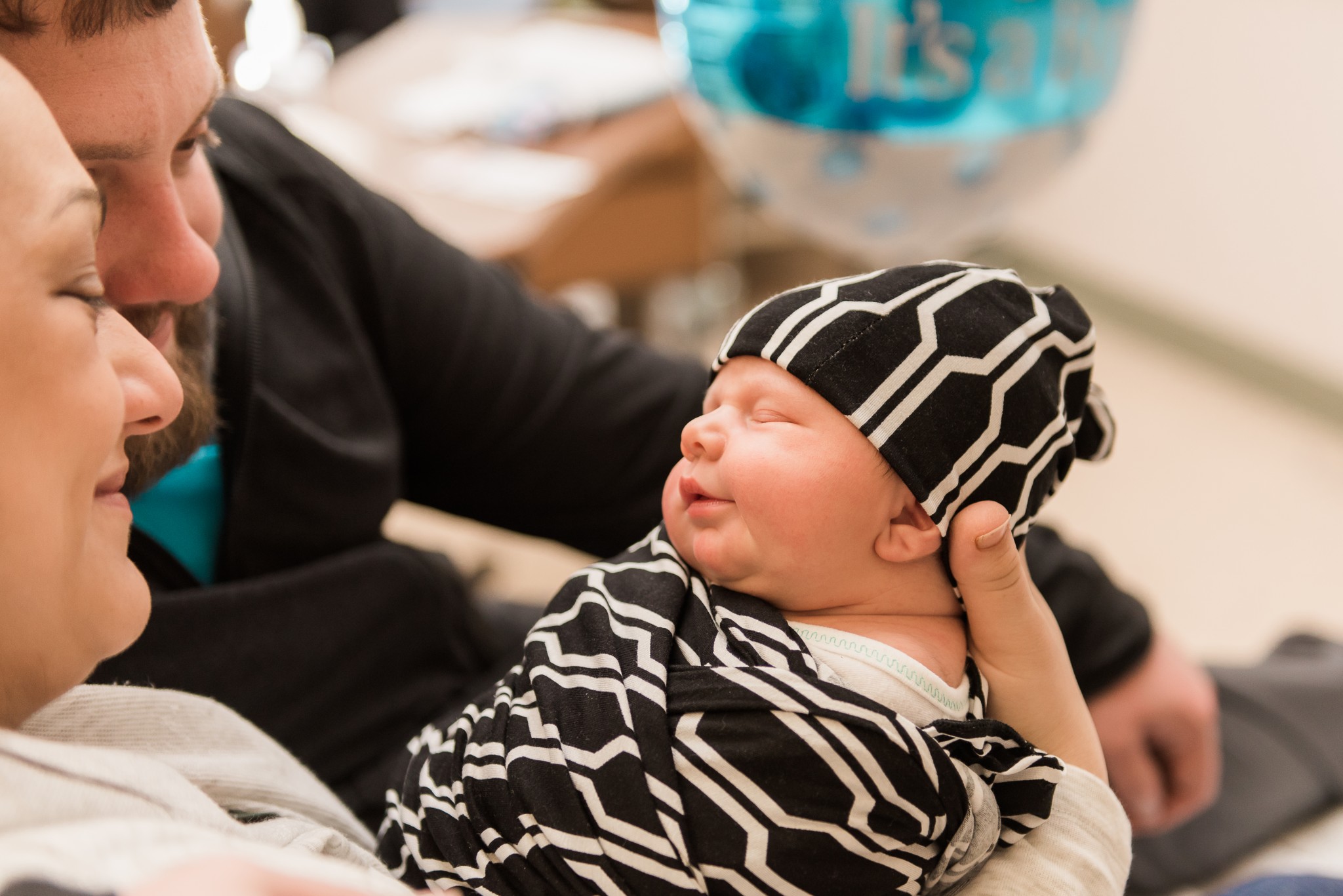A baby boy wearing a black and white geometric swaddle is held by his parents as they look on during their photography session in a Pittsburgh hospital.