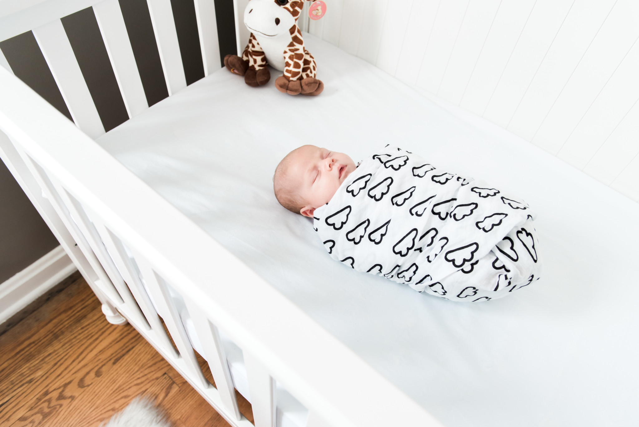 Baby boy wrapped in black and white swaddle sleeps in his white crib with a stuffed giraffe during his in home lifestyle photography session in Pittsburgh and Cranberry Twp PA 1