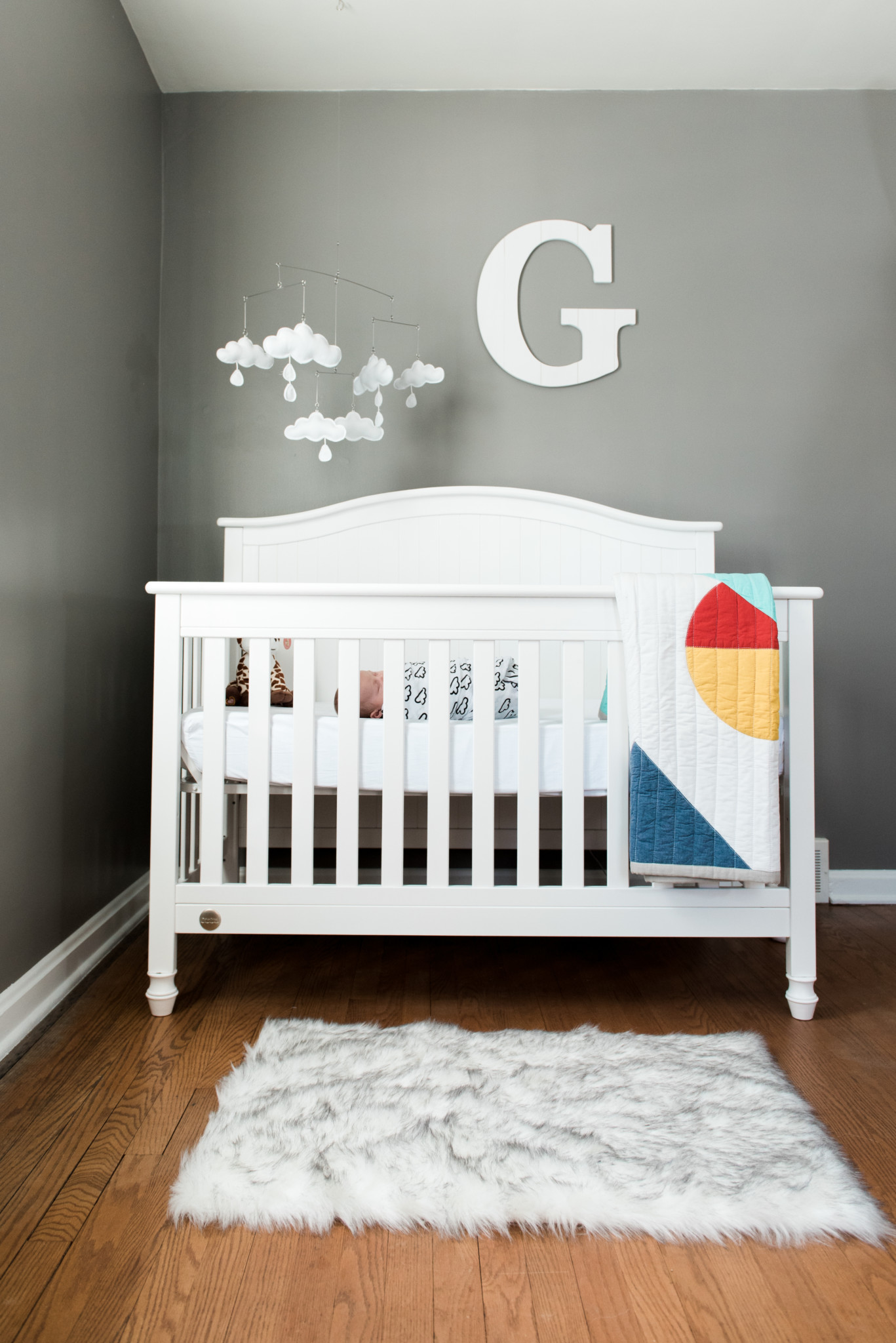 Baby boy wrapped in a white swaddle lays sleeping in his white crib. Above the crib on the wall is a large letter G and a cloud mobile. On the floor lays a white fur rug and on the crib hangs a colorful quilt for this in home newborn lifestyle photography session in Pittsburgh, PA